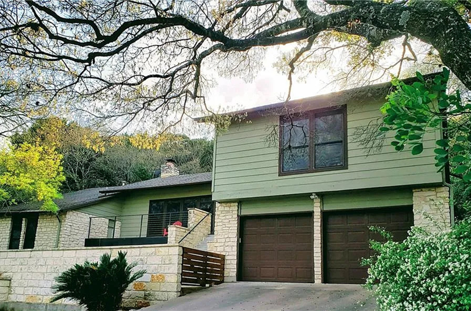 Front exterior before an Austin remodel, showing green siding, white stone, brown garage doors, a wood entry landing and mature trees.