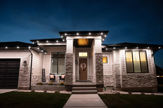 Title: JellyFish Lighting Jelly Lights - Description: White JellyFish eave lights outlining the roofline of a house with a covered entry, white siding and stone columns at dusk.